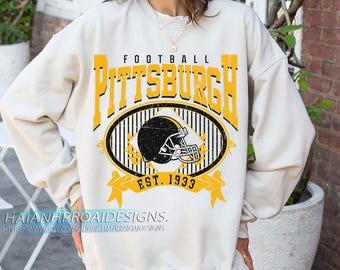 May include: Cream sweatshirt with a yellow and black graphic. The graphic features the word "PITTSBURGH" in yellow block letters above an oval with a football helmet and the text "EST. 1933".