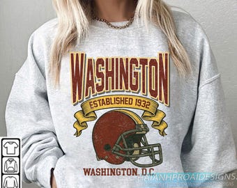 Op de afbeelding: Een grijze sweatshirt met een retro-ontwerp. Het ontwerp bevat het woord "WASHINGTON" in bordeauxrood en goud, de tekst "ESTABLISHED 1932" en een bordeauxrode voetbalhelm. De tekst "WASHINGTON, D.C." is ook inbegrepen.