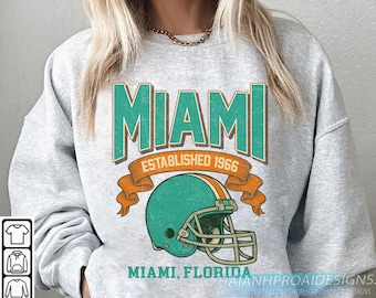 Könnte beinhalten: Ein hellgraues Sweatshirt mit einer türkis-orangenen Grafik eines Football-Helms und dem Wort "MIAMI" in türkisfarbenen Blockbuchstaben. Der Text "ESTABLISHED 1966" und "MIAMI, FLORIDA" sind ebenfalls abgebildet.