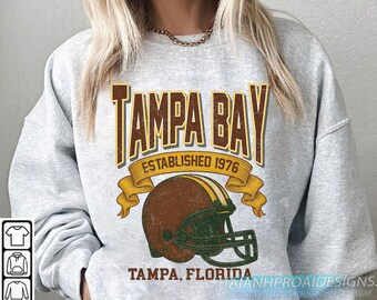 Puede incluir: Sudadera gris claro con un gráfico desgastado. Presenta un casco de fútbol americano marrón y dorado y las palabras "TAMPA BAY ESTABLISHED 1976". Debajo, se lee "TAMPA, FLORIDA".