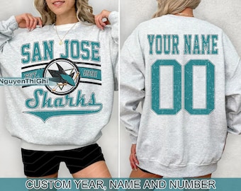May include: A light gray sweatshirt with the text "SAN JOSE Sharks" in teal and white. The front features a shark logo and the year "1991." The back displays "YOUR NAME" and the number "00" in teal. The bottom of the image reads "CUSTOM YEAR, NAME AND NUMBER."