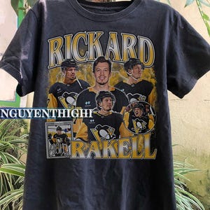 May include: Black t-shirt featuring a graphic print of hockey player Rickard Rakell wearing a Pittsburgh Penguins jersey. The graphic includes the text "Rickard" and "Rakell" in yellow.