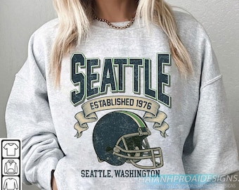 May include: A light grey sweatshirt with a distressed green and white "SEATTLE" graphic, and "ESTABLISHED 1976" below. A football helmet graphic is centered, with "SEATTLE, WASHINGTON" below. The design has a vintage feel.