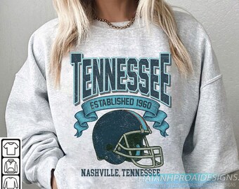 Op de afbeelding: Lichtgrijze sweatshirt met een distressed ontwerp in blauwgroen. Het ontwerp bevat het woord "TENNESSEE", de tekst "ESTABLISHED 1960" en een afbeelding van een footballhelm. De tekst "NASHVILLE, TENNESSEE" staat eronder.