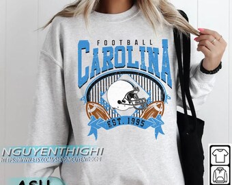 Może przedstawiać: Jasnoszara bluza z nadrukiem futbolu amerykańskiego Carolina. Projekt zawiera napis "CAROLINA" w jasnoniebieskim kolorze, kask futbolowy i dwie piłki do futbolu. Widoczny jest również napis "EST. 1995". Bluza ma okrągły dekolt.
