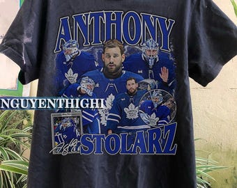 Puede incluir: Camiseta gris oscuro con un collage de un jugador de hockey con uniforme azul y blanco. El nombre "ANTHONY STOLARZ" está impreso en azul y blanco, con el logotipo del equipo visible.