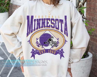 Puede incluir: Sudadera de color crema con un gráfico de fútbol morado y dorado. El gráfico presenta la palabra "MINNESOTA" en letras moradas grandes, un óvalo con un casco de fútbol y el texto "EST. 1968".