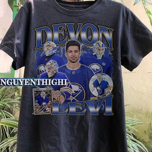 May include: Black t-shirt with a graphic print of a hockey player, Devon Levi, with the text "Devon Levi" in yellow and blue letters. The graphic also includes a hockey stick and a hockey puck.