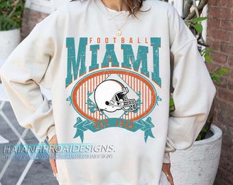 Könnte beinhalten: Cremefarbenes Sweatshirt mit einem verblassten türkis-orangenen Aufdruck. Er lautet "FOOTBALL MIAMI" über einem ovalen Design mit einem Football-Helm. Der Text "EST. 1975" steht darunter. Der Shop-Name und die URL stehen unten.