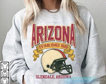 Op de afbeelding: Een lichtgrijs sweatshirt met een vintage-stijl grafische afbeelding. De afbeelding toont het woord "ARIZONA" in bordeauxrood, met "ESTABLISHED 1920" eronder. Een afbeelding van een voetbalhelm is inbegrepen. De tekst "GLENDALE, ARIZONA" staat eronder.