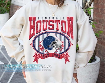 Op de afbeelding: Crèmekleurige sweatshirt met een verweerde rode en blauwe afbeelding. De afbeelding toont het woord "HOUSTON" boven een ovaal ontwerp met een footballhelm en de tekst "EST. 1999". De sweatshirt heeft lange mouwen.