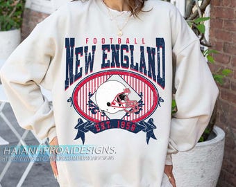 以下が含まれることがあります： クリーム色のスウェットシャツ。デザインは、ネイビーブルーの「FOOTBALL NEW ENGLAND」の文字と、フットボールヘルメットの入った赤と白の楕円で構成されています。「EST. 1959」の文字がバナーに表示されています。