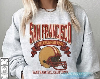 Puede incluir: Una sudadera gris claro con un gráfico de estilo vintage. El diseño incluye las palabras "SAN FRANCISCO" en una fuente roja desgastada, con "ESTABLISHED 1946" debajo. También se presenta un casco de fútbol. Debajo del casco está el texto "SAN FRANCISCO, CALIFORNIA".