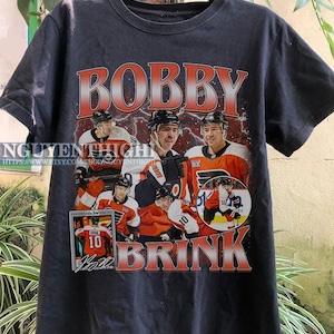 May include: Black t-shirt with a graphic print of hockey player Bobby Brink. The graphic features a collage of Brink in different hockey jerseys, including a Flyers jersey with the number 10. The text "Bobby Brink" is printed in red and white letters above the graphic, and the text "Brink" is printed in red and white letters below the graphic.