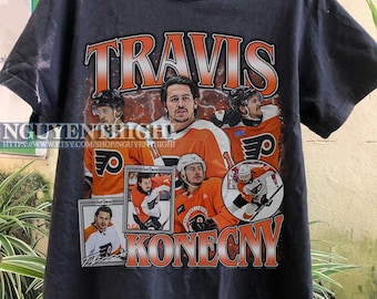 Pode incluir: Camiseta preta com uma estampa gráfica do jogador de hóquei Travis Konecny vestindo uma camisa dos Philadelphia Flyers. O gráfico inclui o texto "Travis Konecny" e imagens de Konecny em diferentes poses.