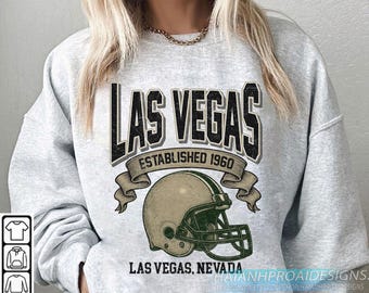 Può includere: Felpa grigio chiaro con grafica invecchiata. Presenta la scritta "LAS VEGAS" sopra un casco da football americano. Sotto, "LAS VEGAS, NEVADA" e "ESTABLISHED 1960". Il design è in nero e verde oliva.