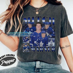 May include: Dark gray t-shirt featuring a graphic of Mitch Marner, a hockey player, in a blue and white uniform. The shirt includes the text "MITCH MARNER" in a blue and white design, with additional images of the player.