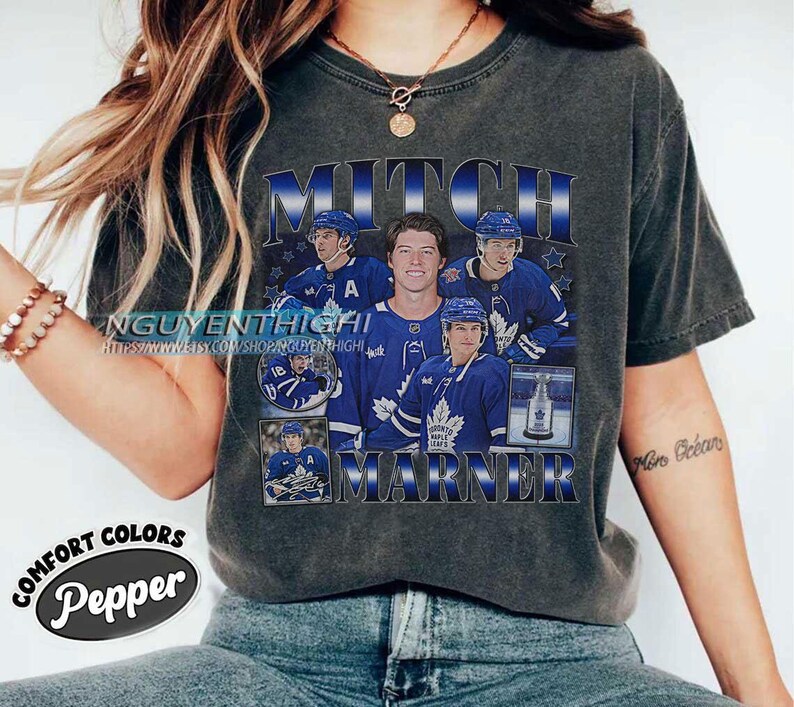 May include: Dark gray t-shirt featuring a graphic of Mitch Marner, a hockey player, in a blue and white uniform. The shirt includes the text "MITCH MARNER" in a blue and white design, with additional images of the player.