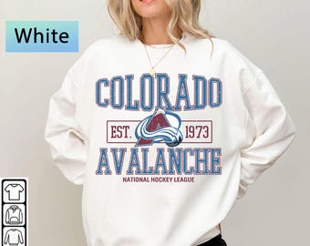 May include: White sweatshirt featuring the text "COLORADO AVALANCHE" in blue and maroon, with the team logo and "EST. 1973". The words "NATIONAL HOCKEY LEAGUE" are printed below the team name. The word "White" is in a blue box.