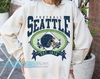 Könnte beinhalten: Cremefarbenes Sweatshirt mit einem Seattle Football-Motiv. Das Design zeigt das Wort "FOOTBALL" über "SEATTLE" in Dunkelblau, mit einem Footballhelm in einem grün-weißen Oval. Der Text "EST. 1976" steht darunter.