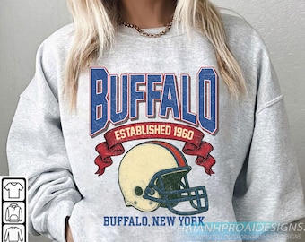 May include: A light grey sweatshirt with a distressed graphic. The graphic features the word "BUFFALO" in blue and red, with "ESTABLISHED 1960" and a football helmet. The text "BUFFALO, NEW YORK" is below the helmet.