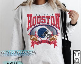 Könnte beinhalten: Ein hellgraues Sweatshirt mit einem Used-Look-Grafikdesign. Das Design zeigt das Wort "FOOTBALL" über "HOUSTON" in Rot, einen blauen Footballhelm und zwei Fußbälle. Der Text "EST. 1999" ist ebenfalls enthalten.