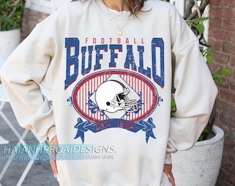 May include: A cream-coloured sweatshirt with a distressed blue and red graphic. The graphic features the word "BUFFALO", a football helmet, and the text "EST. 1960". The sweatshirt has long sleeves and a crew neck.