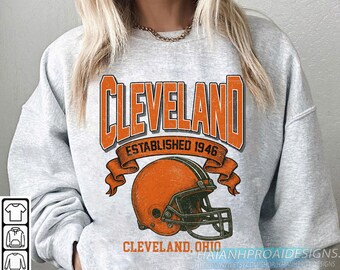 May include: A light gray sweatshirt featuring the word "Cleveland" in orange block letters above an orange football helmet. The text "Established 1946" is above the helmet. The words "Cleveland, Ohio" are below the helmet.