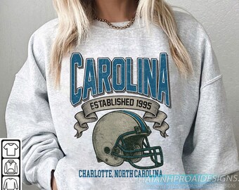 Może przedstawiać: Jasnoszara bluza z nadrukiem w kolorze niebieskim "CAROLINA" w stylu vintage, napisem "ESTABLISHED 1995" i grafiką kasku do futbolu amerykańskiego. Zawiera również tekst "CHARLOTTE, NORTH CAROLINA".
