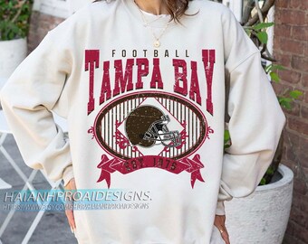 Puede incluir: Sudadera color crema con un gráfico desgastado en burdeos y marrón. El diseño incluye las palabras "FOOTBALL TAMPA BAY" y un casco de fútbol dentro de un marco ovalado. También es visible el texto "EST. 1975".