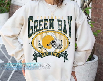 Peut inclure: Sweat-shirt crème avec un graphisme vieilli. Il présente un casque de football à l'intérieur d'un ovale avec les mots "GREEN BAY FOOTBALL" et "EST. 1962". Le design est vert et jaune.