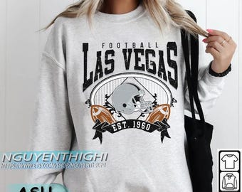 Può includere: Felpa grigio chiaro con grafica invecchiata "LAS VEGAS FOOTBALL", un casco da football e due palloni da football. La grafica include "EST. 1960". La felpa ha anche il testo "NGUYENTHIGHI" e "ASH" stampati.