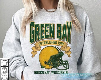 Peut inclure: Un sweat-shirt gris clair avec un design de style vintage. Le design comprend les mots "GREEN BAY" en vert, "ESTABLISHED 1919", un casque de football américain jaune et les mots "GREEN BAY, WISCONSIN".