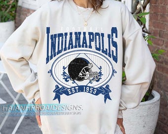 May include: Cream-colored sweatshirt with a blue Indianapolis football design. The design includes the word "Indianapolis" above an oval with a football helmet and the text "EST. 1953" on a banner.