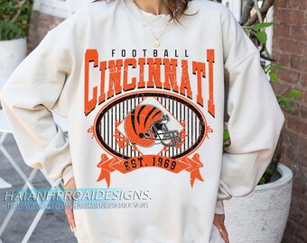 Può includere: Felpa color crema con la scritta "CINCINNATI" in lettere arancioni a blocchi sopra un disegno ovale con un casco da football. Il design include il testo "FOOTBALL" e "EST. 1969".