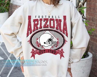 Op de afbeelding: Crèmekleurige sweatshirt met een verweerde bordeauxrode afbeelding. De afbeelding toont het woord "FOOTBALL" boven "ARIZONA" in vette letters, een ovaal met een footballhelm en "EST. 1989" op een banner. De Etsy shopnaam is onderaan zichtbaar.