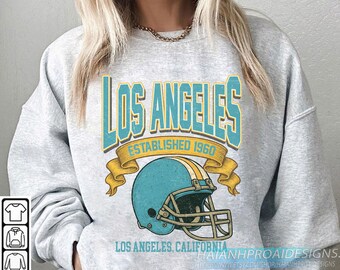 Op de afbeelding: Een lichtgrijs sweatshirt met een turquoise en gele afbeelding. De afbeelding luidt "LOS ANGELES ESTABLISHED 1960" boven een turquoise American footballhelm. De tekst "LOS ANGELES, CALIFORNIA" staat eronder.