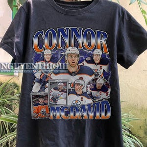 May include: Black t-shirt with a graphic of hockey player Connor McDavid. The graphic features a collage of McDavid's hockey cards and a signature. The text "Connor McDavid" is printed in large, bold letters.