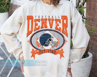 May include: Cream-coloured sweatshirt with the word "DENVER" in orange block letters above an oval design featuring a blue American football helmet. The text "EST. 1969" is below the oval. The sweatshirt has long sleeves.