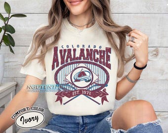 May include: A white t-shirt with a burgundy and blue graphic of the Colorado Avalanche logo. The logo features a mountain range and a hockey stick with the text "Colorado Avalanche Est. 1972".