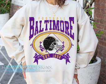 Puede incluir: Sudadera color crema con un gráfico morado y dorado. El gráfico presenta la palabra "BALTIMORE" sobre un casco de fútbol dentro de un óvalo, con "EST. 1996" debajo. La sudadera tiene mangas largas.