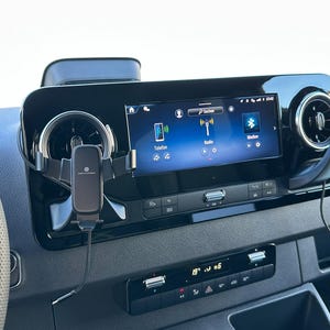 K&ouml;nnte beinhalten: Fahrzeug-Armaturenbrett mit zentralem Infotainment-Bildschirm, der Symbole f&uuml;r Telefon, Radio und Bluetooth anzeigt. Eine schwarze Telefonhalterung ist an der L&uuml;ftungs&ouml;ffnung befestigt. Das Armaturenbrett ist schwarz mit silbernen Akzenten.