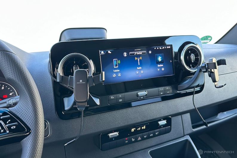 K&ouml;nnte beinhalten: Fahrzeug-Armaturenbrett mit zentralem Infotainment-Bildschirm, der Symbole f&uuml;r Telefon, Radio und Bluetooth anzeigt. Eine schwarze Telefonhalterung ist an der L&uuml;ftungs&ouml;ffnung befestigt. Das Armaturenbrett ist schwarz mit silbernen Akzenten.