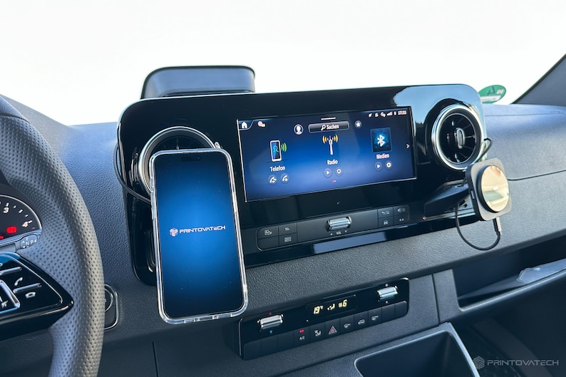 K&ouml;nnte beinhalten: Ein Armaturenbrett im Auto mit einem Smartphone in einer transparenten H&uuml;lle, das am Armaturenbrett befestigt ist. Das Telefon zeigt das Printovatech-Logo an. Das Armaturenbrett hat ein digitales Display mit Radio- und Telefonoptionen.
