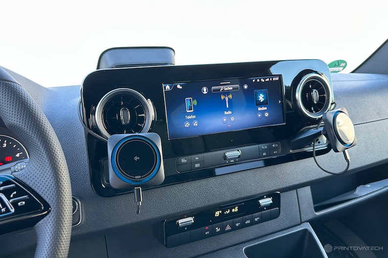 K&ouml;nnte beinhalten: Fahrzeug-Armaturenbrett mit zentralem Infotainment-Bildschirm, der Telefon-, Radio- und Bluetooth-Symbole anzeigt. Ein kabelloses Ladepad und eine Telefonhalterung sind am Armaturenbrett montiert. Das Armaturenbrett ist schwarz mit silbernen Akzenten.