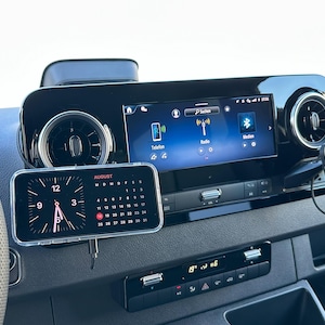 K&ouml;nnte beinhalten: Ein Fahrzeug-Armaturenbrett mit einem Smartphone, das eine Uhr und einen Kalender f&uuml;r den 12. August anzeigt. Das Armaturenbrett enth&auml;lt auch ein digitales Display mit Radio- und Bluetooth-Optionen sowie L&uuml;ftungsschlitze und Bedienelemente.