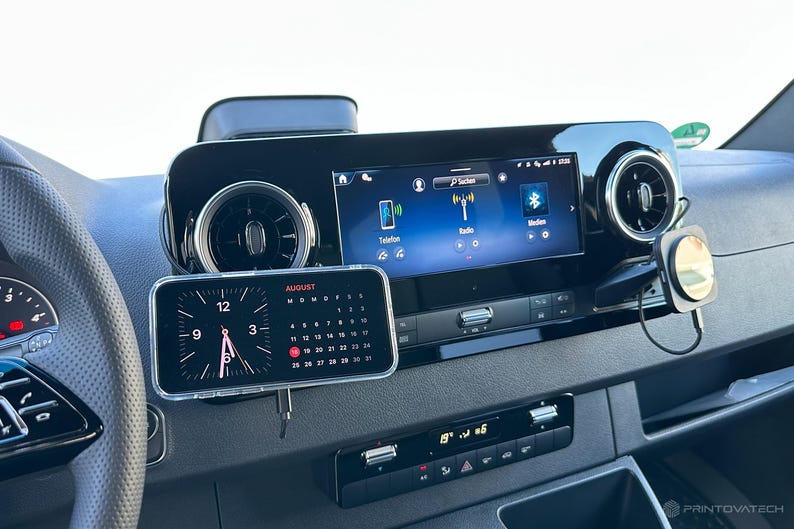 K&ouml;nnte beinhalten: Ein Fahrzeug-Armaturenbrett mit einem Smartphone, das eine Uhr und einen Kalender f&uuml;r den 12. August anzeigt. Das Armaturenbrett enth&auml;lt auch ein digitales Display mit Radio- und Bluetooth-Optionen sowie L&uuml;ftungsschlitze und Bedienelemente.