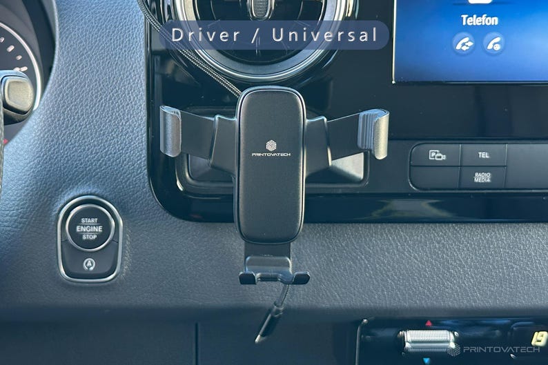 K&ouml;nnte beinhalten: Ein schwarzer Printovatech-Telefonhalter, der in einem Auto montiert ist. Der Halter ist am Armaturenbrett befestigt und verf&uuml;gt &uuml;ber verstellbare Arme zur Sicherung eines Telefons. Das Armaturenbrett des Autos umfasst eine Start/Stopp-Taste und einen Bildschirm.
