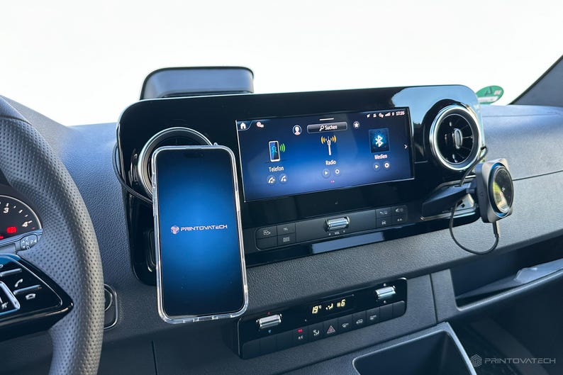 K&ouml;nnte beinhalten: Ein Armaturenbrett im Auto mit einem Smartphone in einer transparenten H&uuml;lle, montiert neben einem digitalen Display. Das Telefon zeigt das Printovatech-Logo auf blauem Farbverlauf. Das Armaturenbrett hat auch L&uuml;ftungsd&uuml;sen und Klimaanlagen.