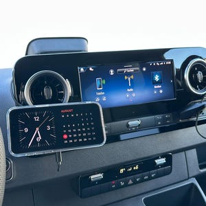 K&ouml;nnte beinhalten: Ein Armaturenbrett im Auto mit einem digitalen Display, das Radio- und Telefonoptionen anzeigt. Ein Smartphone zeigt eine Uhr und einen Kalender f&uuml;r August an. Ein kleiner Lautsprecher ist am Armaturenbrett montiert. Das Armaturenbrett ist schwarz mit silbernen Akzenten.